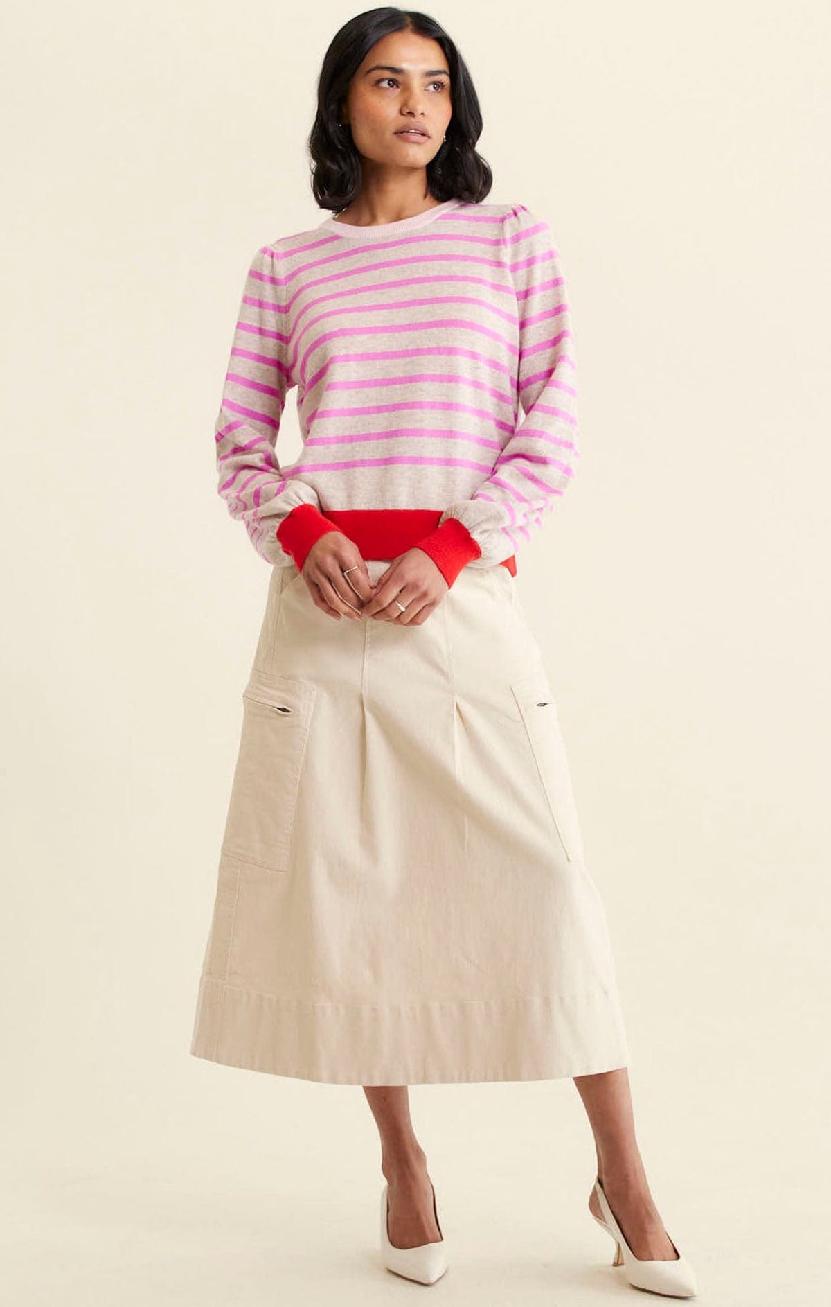 A woman wears the Alessandra CASHMERE BLEND STRIPE KNIT IN PINK— a long-sleeved pink and light gray striped sweater with red cuffs and waistband—paired with a beige A-line skirt and white heels, set against a plain light background.