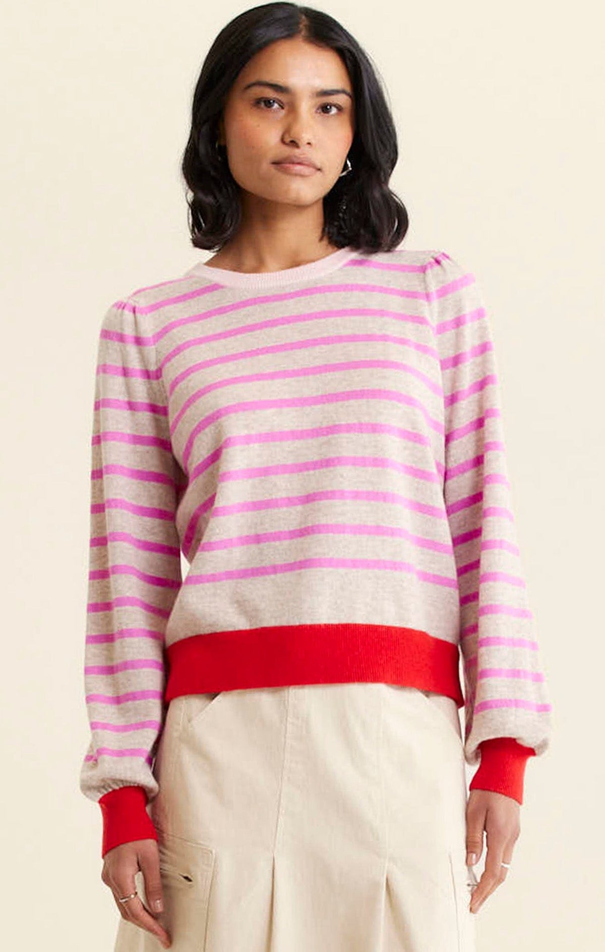 A woman wears the Alessandra CASHMERE BLEND STRIPE KNIT IN PINK— a beige, long-sleeve sweater with pink stripes and red cuffs and hem— paired with a light skirt, standing against a plain beige background.