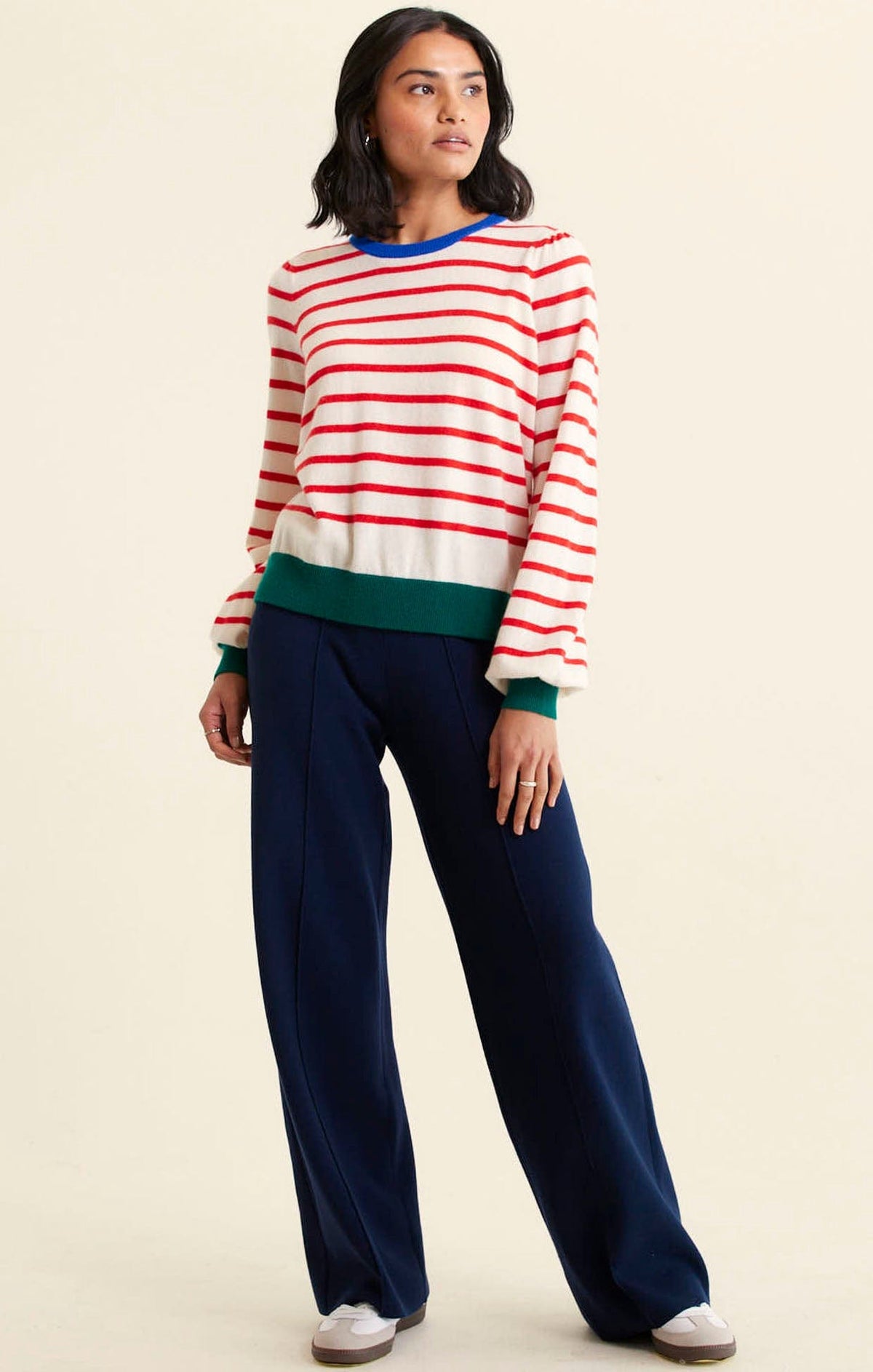 A woman wears the Alessandra CASHMERE BLEND STRIPE KNIT IN RED—white with red stripes, blue collar, green cuffs and hem—with navy wide-leg trousers and white sneakers, gazing off to the side against a plain beige background.