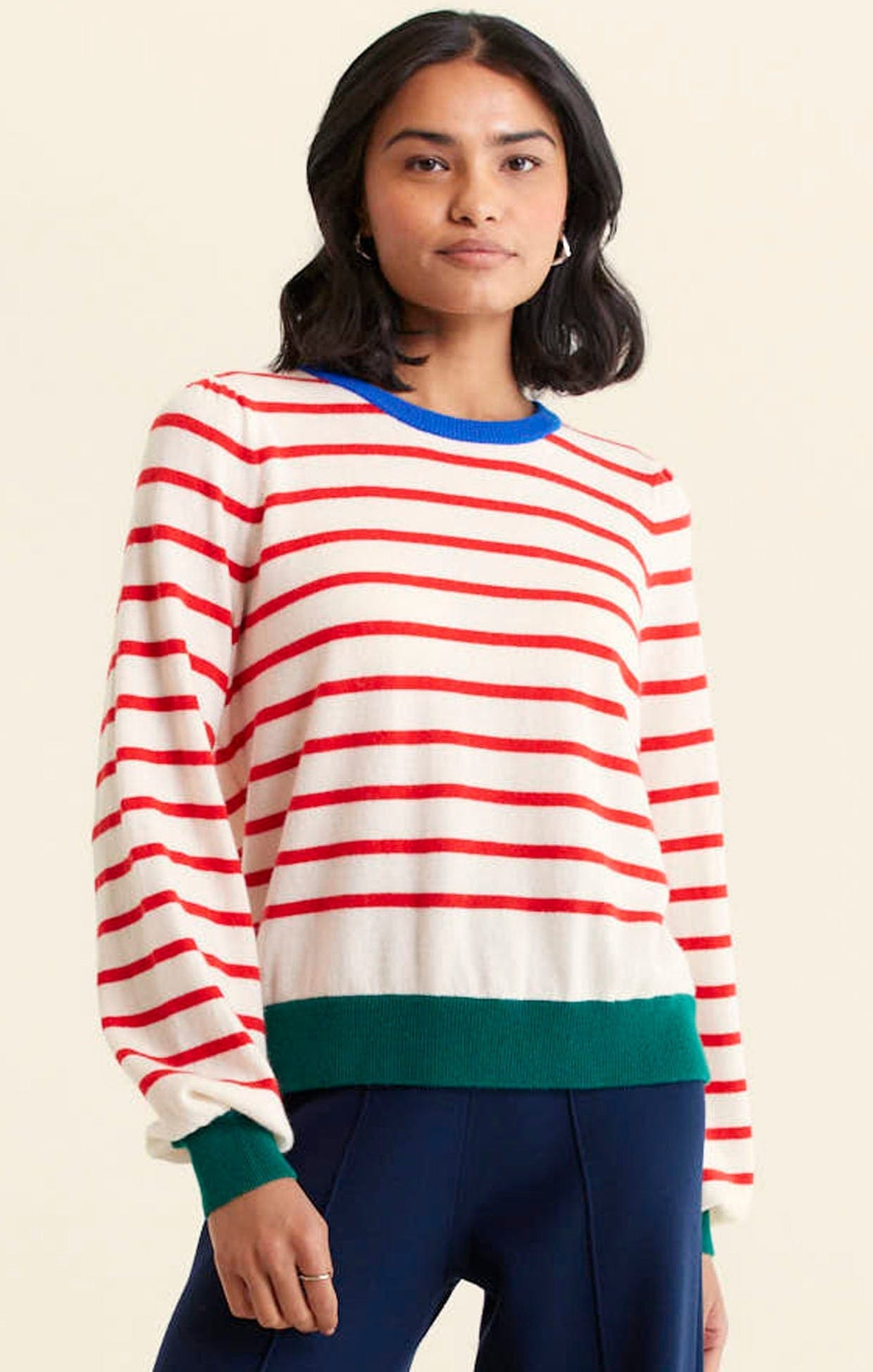 A woman with medium-length dark hair wears the Alessandra CASHMERE BLEND STRIPE KNIT IN RED—featuring a cream base, red stripes, blue collar, and green cuffs and hem—with dark blue pants against a plain light background.