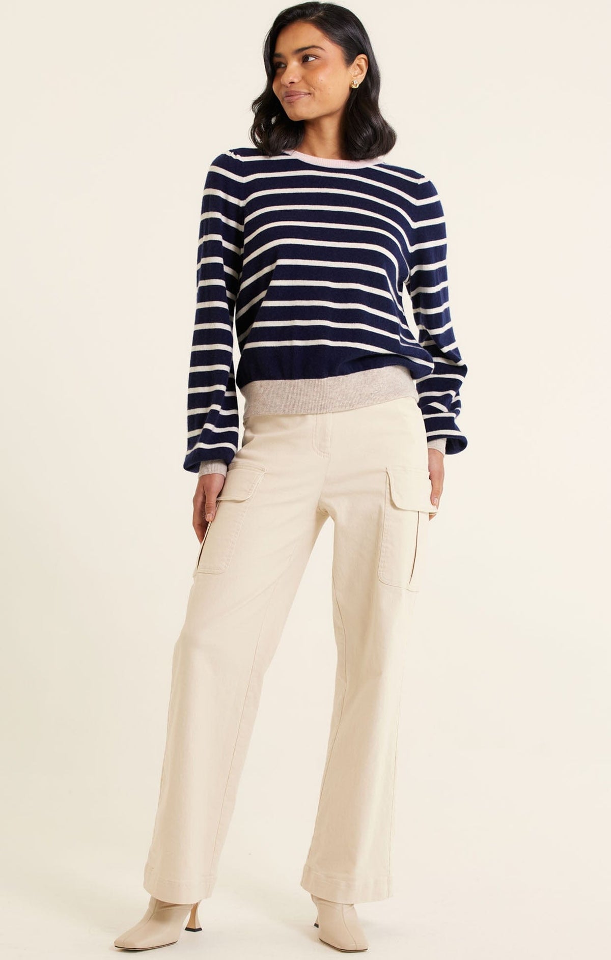 A woman smiles, wearing the Alessandra CASHMERE BLEND STRIPE KNIT IN NAVY, light beige wide-leg cargo pants with side pockets, and beige heeled ankle boots against a plain off-white background.