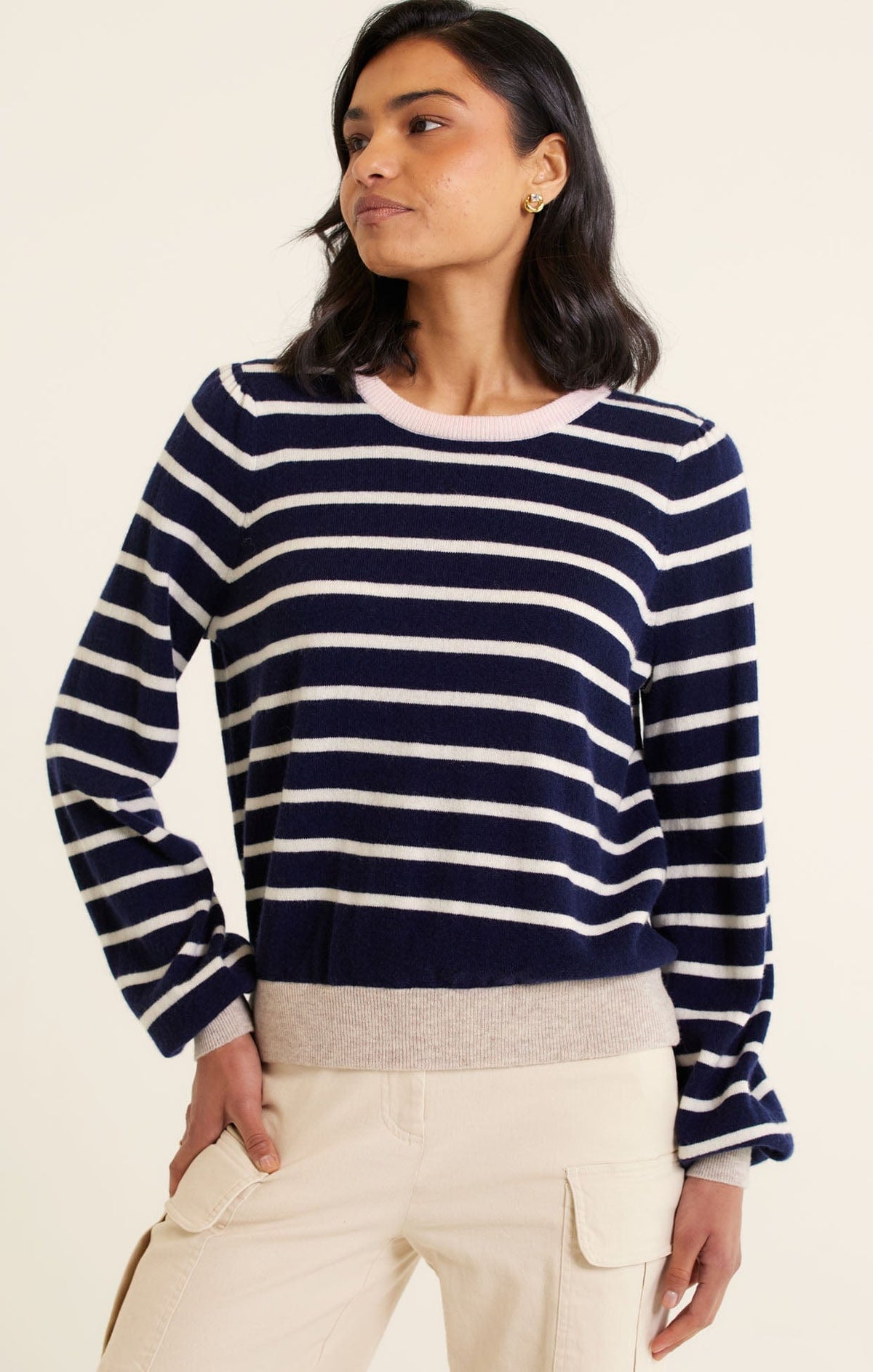 A woman with wavy dark hair models the Alessandra CASHMERE BLEND STRIPE KNIT IN NAVY—navy and white striped with light ribbed cuffs and hem—paired with beige cargo pants, standing against a plain light background.